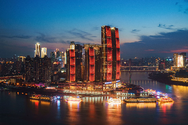 Vista aérea nocturna de los rascacielos iluminados de Raffles City en Chongqing y barcos turísticos en el río. Explora el lado más moderno del país con nuestra agencia de viajes a China a la medida.