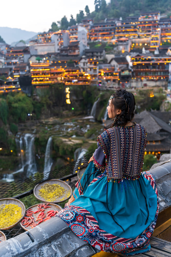 Mujer con vestimenta tradicional contemplando un pueblo antiguo iluminado con cascadas en China. Diseña tus viajes personalizados a China y descubre destinos culturales auténticos con Destino China