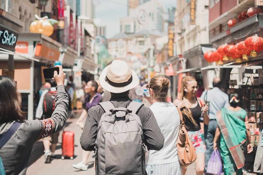 Viajero con mochila y sombrero caminando por una concurrida calle comercial con linternas rojas. Explora los mercados tradicionales con nuestra agencia de viajes a China a la medida.
